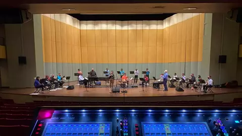 A large ensemble of musicians rehearses on stage, with a sound mixing console in the foreground displaying various controls and settings.