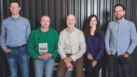 Five individuals pose together, with one holding a book titled "UR 100." They are dressed casually and stand against a dark, textured background.