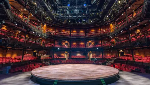 A circular stage is centered in a modern theater, surrounded by tiered seating and overhead lighting fixtures.