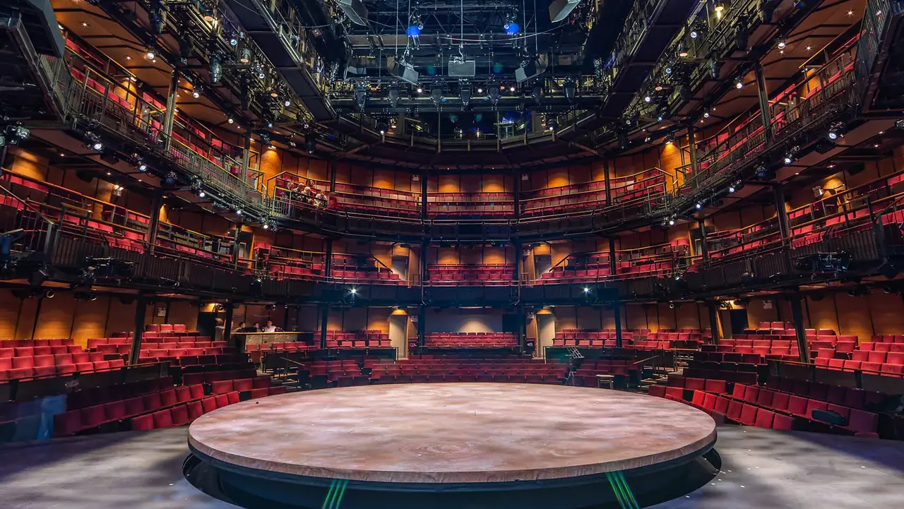 A circular stage is centered in a modern theater, surrounded by tiered seating and overhead lighting fixtures.