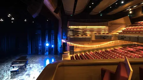 A grand concert hall featuring a grand piano on stage, with rows of red seats and modern lighting design.
