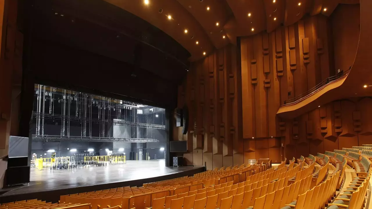 Interior of a theater featuring a spacious stage with rigging and rows of wooden seats, illuminated by overhead lights.