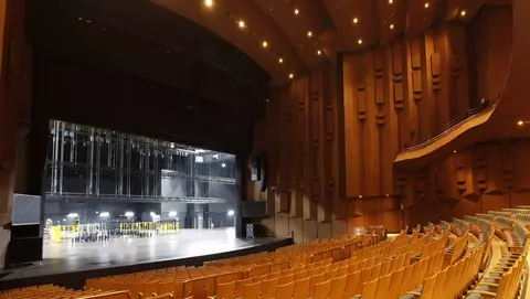 Interior of a theater featuring a spacious stage with rigging and rows of wooden seats, illuminated by overhead lights.