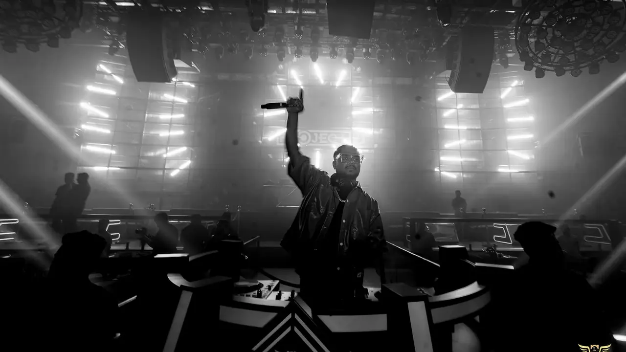 A performer stands on stage with a microphone, raising one hand, surrounded by dim lighting and silhouettes of the audience in a club setting.