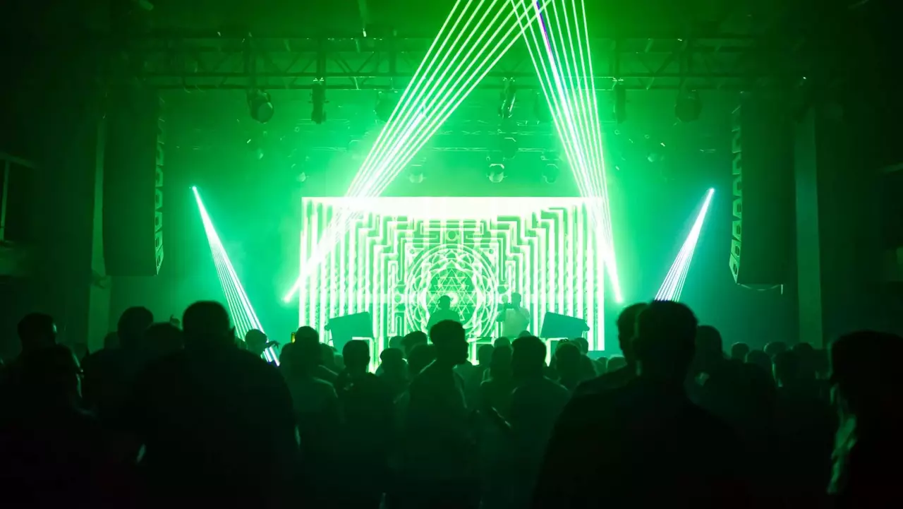 Menschenmenge, die in einem schwach beleuchteten Veranstaltungsort tanzt, umgeben von lebhaften grünen Laserlichtern und einer geometrischen visuellen Darstellung auf der Bühne.