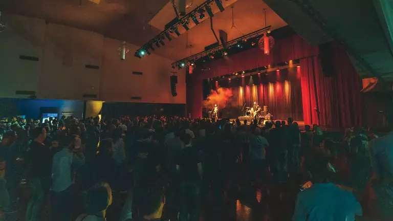 A crowd watches a band perform on stage, illuminated by colorful lights in a large indoor venue with red curtains.