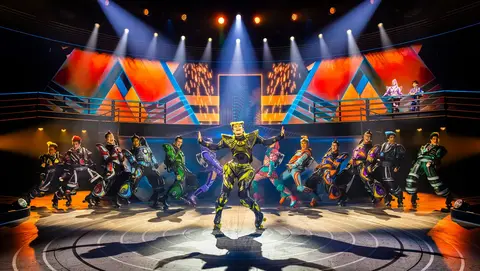 A group of dancers in colorful costumes performs on stage, with a central performer in a striking yellow outfit, surrounded by vibrant lighting and geometric backdrops.
