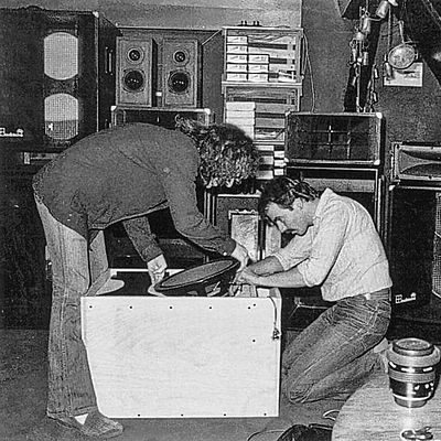 Two individuals work together on assembling audio equipment in a room filled with speakers and recording materials.