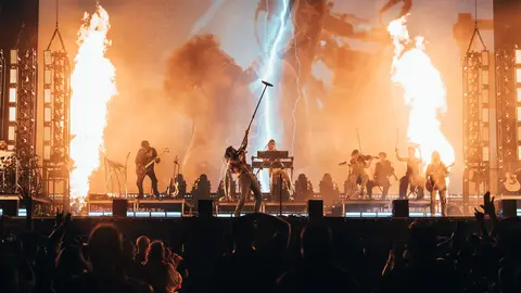 A live music performance featuring multiple musicians on stage, with dramatic lighting, flames, and a large screen displaying visuals in the background.