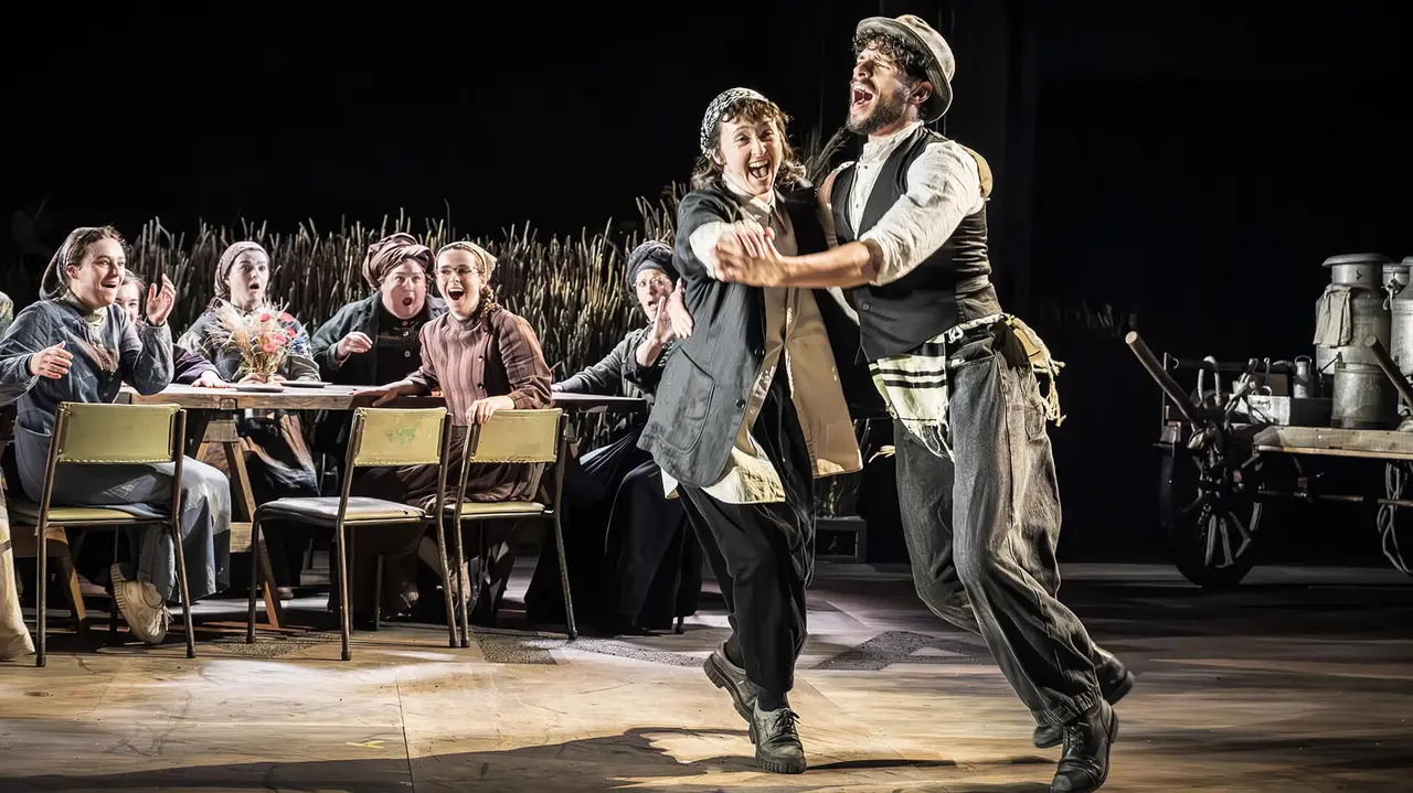 Two performers dance joyfully in a theatrical setting, while a group of seated characters observes, creating a lively scene.
