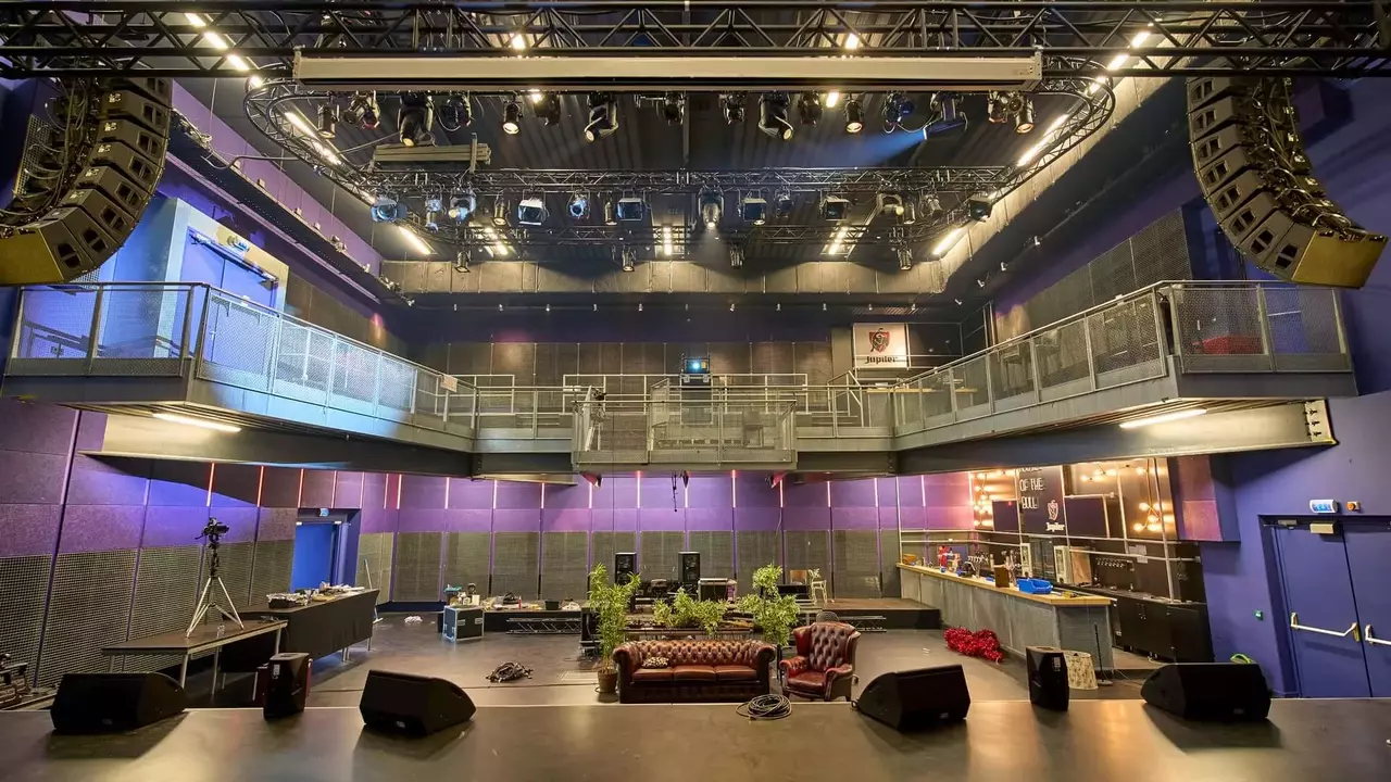 A spacious performance venue featuring a stage with sound equipment, seating areas, and multiple levels, illuminated by stage lights.