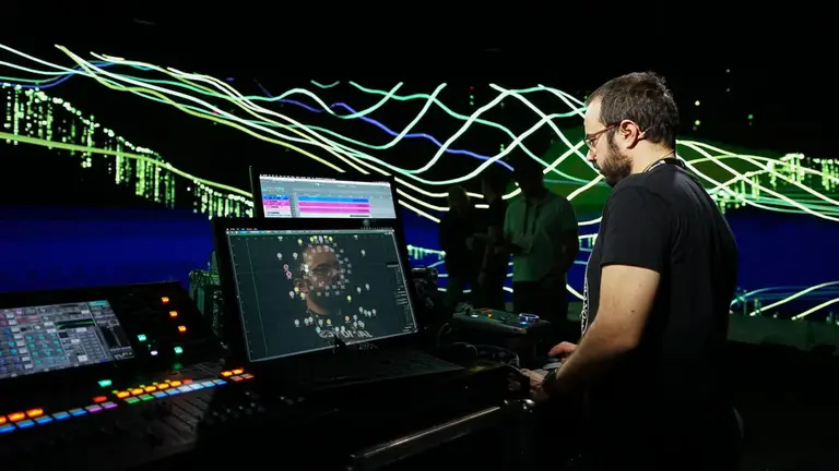 A man operates a digital audio workstation with colorful visualizations in the background, surrounded by equipment and other individuals.