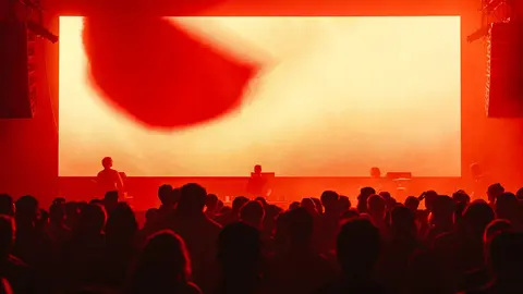 Crowd at a concert with silhouetted performers in front of a large red screen displaying abstract visuals.