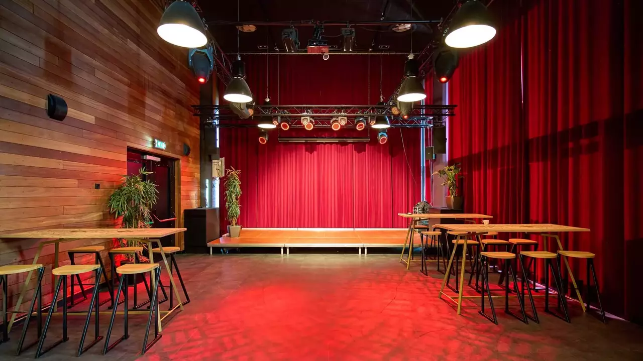 Empty performance space with red curtains, wooden walls, and tall tables surrounded by stools, illuminated by overhead lights.