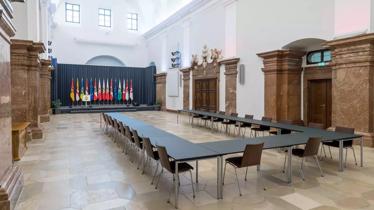 Ein großer Konferenzraum mit einem langen Tisch und Stühlen, im Hintergrund dekoriert mit verschiedenen Nationalflaggen.