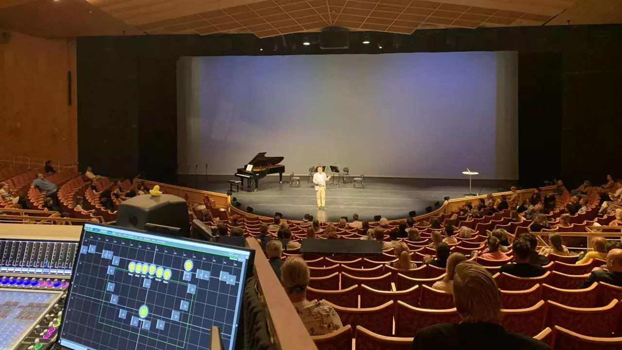 Ein Darsteller steht auf der Bühne in einem Theater vor einem Konzertflügel und leeren Stühlen, während das Publikum aus dem Zuschauerbereich zuschaut.