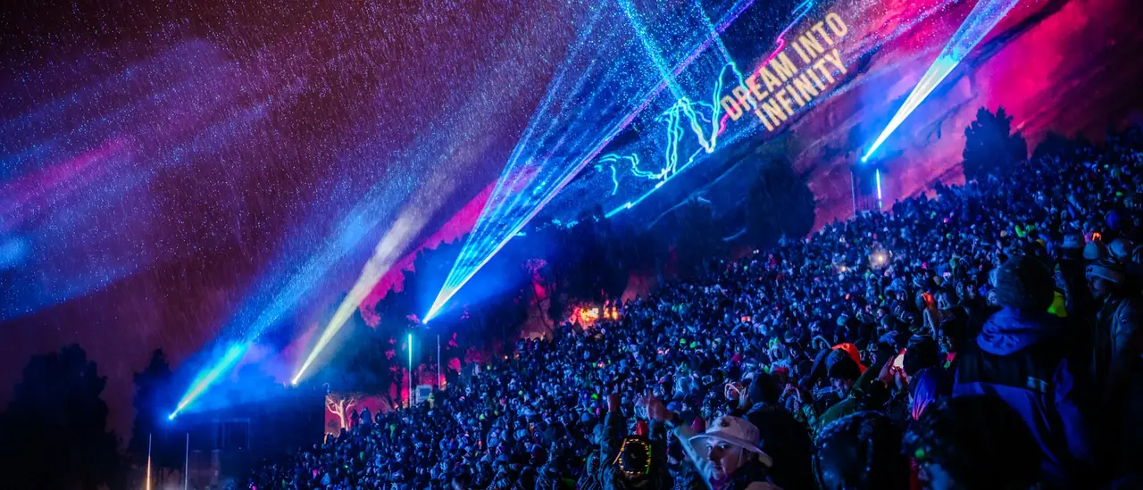 A large crowd gathers under colorful laser lights and snow, with a projection reading "DREAM INTO INFINITY" on a nearby surface.