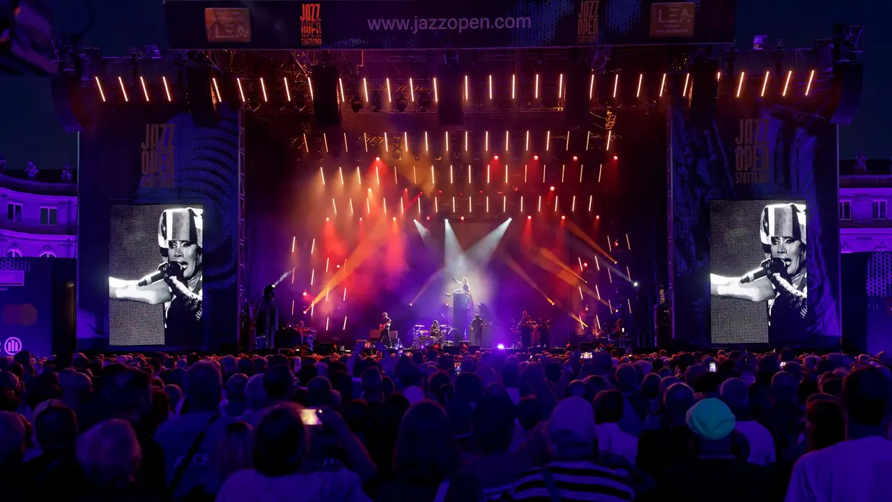 A large crowd watches a live performance on stage, illuminated by colorful lights, with a large screen displaying a musician.