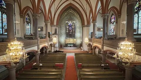Interior view of a church featuring ornate chandeliers, wooden pews, and stained glass windows, with an altar at the front.