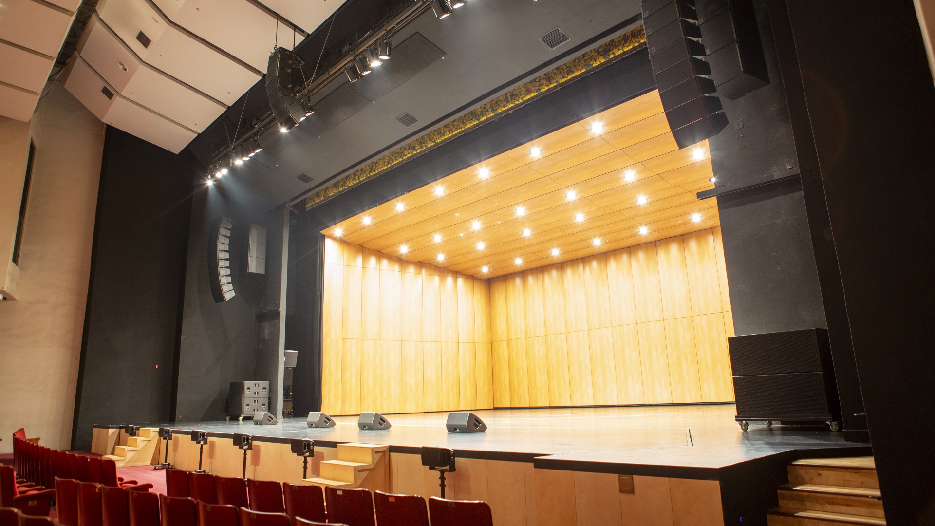 Empty auditorium stage with wooden paneling and overhead lights, featuring sound equipment and red seating in the foreground.